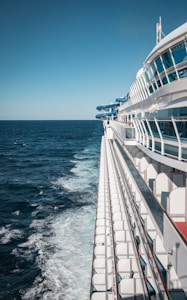 A large cruise ship is navigating through the ocean with its deck and railings prominently visible. The ship's white structure contrasts with the deep blue of the sea. Onboard amenities like a waterslide are visible in the background. The sky is clear and bright, indicating pleasant weather.