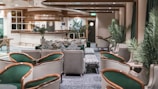 A stylish and elegant lounge area features plush seating with a mix of green and gray armchairs. Zebra-patterned cushions and footrests add a playful touch to the sophisticated decor. The space includes potted plants, enhancing the fresh and modern atmosphere. A polished wooden bar with a circular design and ambient lighting is positioned at the back, creating a welcoming focal point.