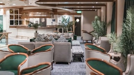 A stylish and elegant lounge area features plush seating with a mix of green and gray armchairs. Zebra-patterned cushions and footrests add a playful touch to the sophisticated decor. The space includes potted plants, enhancing the fresh and modern atmosphere. A polished wooden bar with a circular design and ambient lighting is positioned at the back, creating a welcoming focal point.