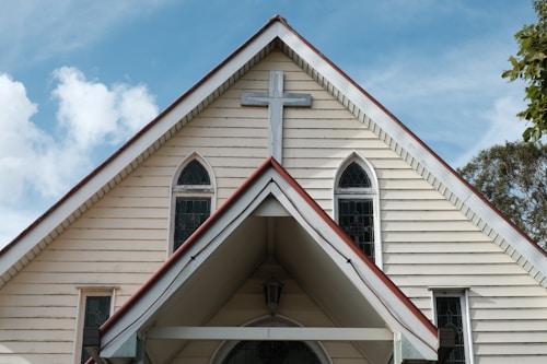 A church facade with a steeply pitched roof featuring a large cross at the apex. The structure is made of light-colored wood with vertical paneling and has three arched windows with dark trim. The sky is partly cloudy, and a few trees can be seen in the background.
