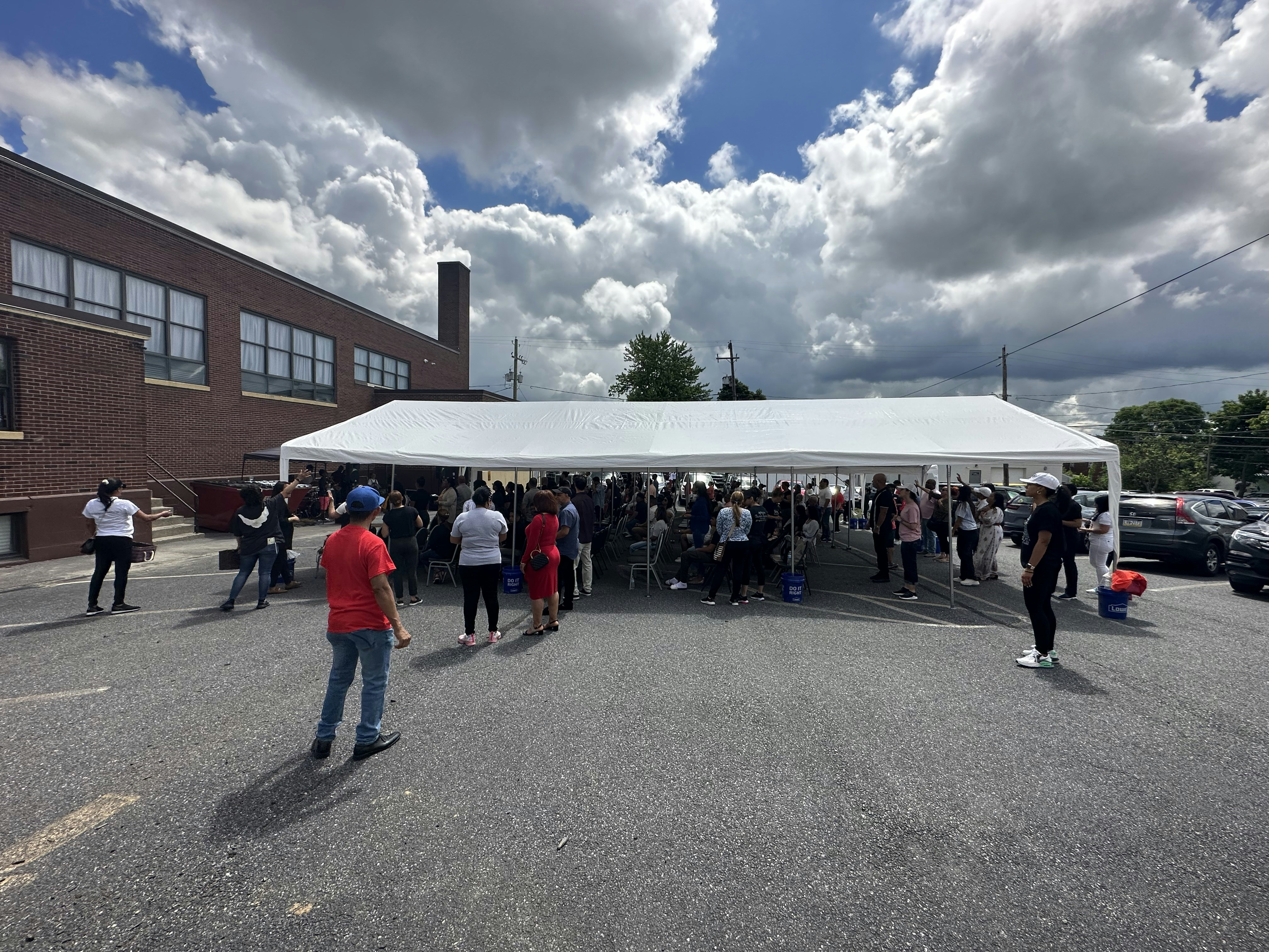 A large group of people gather under a white outdoor tent in a parking lot next to a brick building. The sky is partly cloudy, and several individuals stand around the tent, while others are inside. Cars are parked nearby, and some people are looking down at their phones. The event appears to be social or community-oriented.
