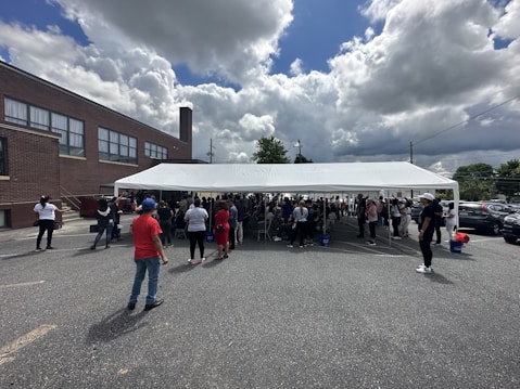 A large group of people gather under a white outdoor tent in a parking lot next to a brick building. The sky is partly cloudy, and several individuals stand around the tent, while others are inside. Cars are parked nearby, and some people are looking down at their phones. The event appears to be social or community-oriented.