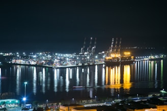 A brightly lit port at night with large cranes and numerous shipping containers. The reflections of the lights in the calm water create a striking visual effect. A large ship is docked at the port, illuminated with an intense golden glow, casting reflections onto the water. Various industrial buildings and infrastructure are visible along the waterfront.