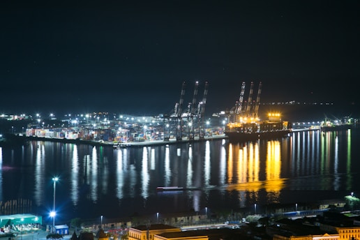 A brightly lit port at night with large cranes and numerous shipping containers. The reflections of the lights in the calm water create a striking visual effect. A large ship is docked at the port, illuminated with an intense golden glow, casting reflections onto the water. Various industrial buildings and infrastructure are visible along the waterfront.