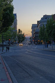 A peaceful city street at dusk with streetlights and people walking safely.