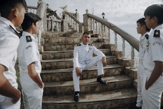 A group of five men in formal white uniforms is gathered around a staircase made of stone. The man in the center is sitting on one of the steps, while the others stand around him, engaging in conversation. The scene is outdoors, with a backdrop of a cloudy sky and a railing adorned with small lanterns.