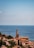 A picturesque coastal town with terracotta-roofed buildings nestled alongside the Mediterranean Sea. A prominent church tower stands tall amidst the buildings, surrounded by lush greenery. The expansive sea stretches out to the horizon, with a few sailboats visible in the distance under a clear blue sky.