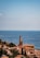 A picturesque coastal town with terracotta-roofed buildings nestled alongside the Mediterranean Sea. A prominent church tower stands tall amidst the buildings, surrounded by lush greenery. The expansive sea stretches out to the horizon, with a few sailboats visible in the distance under a clear blue sky.