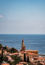 A picturesque coastal town with terracotta-roofed buildings nestled alongside the Mediterranean Sea. A prominent church tower stands tall amidst the buildings, surrounded by lush greenery. The expansive sea stretches out to the horizon, with a few sailboats visible in the distance under a clear blue sky.