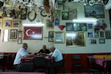 A group of three men sit at a table in a room filled with framed pictures and clocks on the walls. The Turkish flag is prominently displayed. The ceiling is adorned with various lighting fixtures, and the atmosphere is reminiscent of a traditional cafe or gathering space.