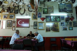 A group of three men sit at a table in a room filled with framed pictures and clocks on the walls. The Turkish flag is prominently displayed. The ceiling is adorned with various lighting fixtures, and the atmosphere is reminiscent of a traditional cafe or gathering space.
