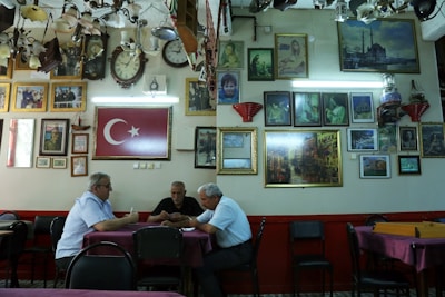 A group of three men sit at a table in a room filled with framed pictures and clocks on the walls. The Turkish flag is prominently displayed. The ceiling is adorned with various lighting fixtures, and the atmosphere is reminiscent of a traditional cafe or gathering space.