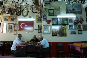 A group of three men sit at a table in a room filled with framed pictures and clocks on the walls. The Turkish flag is prominently displayed. The ceiling is adorned with various lighting fixtures, and the atmosphere is reminiscent of a traditional cafe or gathering space.