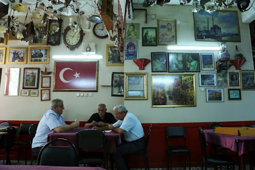 A group of three men sit at a table in a room filled with framed pictures and clocks on the walls. The Turkish flag is prominently displayed. The ceiling is adorned with various lighting fixtures, and the atmosphere is reminiscent of a traditional cafe or gathering space.