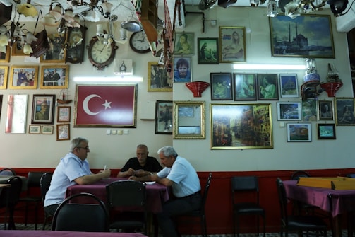 A group of three men sit at a table in a room filled with framed pictures and clocks on the walls. The Turkish flag is prominently displayed. The ceiling is adorned with various lighting fixtures, and the atmosphere is reminiscent of a traditional cafe or gathering space.