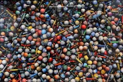 A collection of vibrantly colored balls and variously shaped pin-like objects arranged closely together, creating a visually stimulating pattern. The items display a range of colors including red, blue, yellow, green, and orange.
