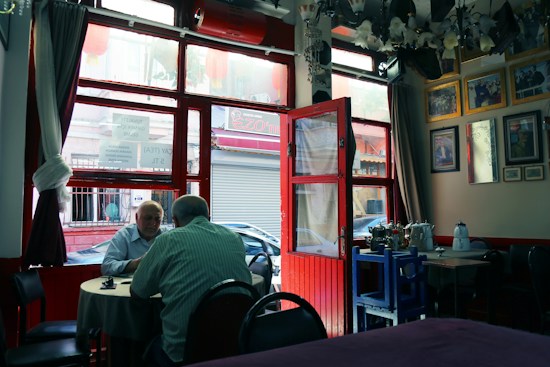 A cozy indoor cafe setting with two elderly men sitting at a round table, engaged in conversation. The room features large windows with a view of the street outside, and the decor includes framed pictures and curtains. There are tables and chairs, as well as teapots and other dining items on nearby surfaces.