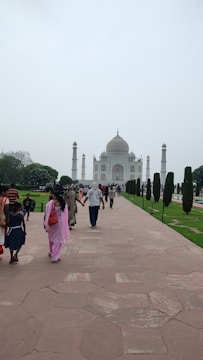 A path leading towards a grand white marble mausoleum with a large dome and four minarets. The surrounding area is lush with well-manicured green lawns and lined with tall, symmetrical trees. People are walking along the path, some in colorful traditional clothing.