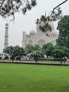 A historic white marble mausoleum with a large dome, surrounded by lush green grass and trees. The scene includes a minaret on the left and is framed by leafy branches. There are people visible in the background, visiting the site, adding a sense of scale.