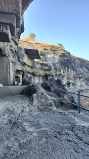 The image shows a series of ancient rock-cut caves with intricately carved facades set against a backdrop of rocky cliffs. The area has a rugged terrain with a clear blue sky above and sparse vegetation in the distance. Visitors can be seen walking along the path near the caves.