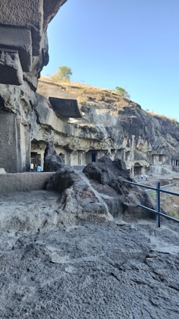 The image shows a series of ancient rock-cut caves with intricately carved facades set against a backdrop of rocky cliffs. The area has a rugged terrain with a clear blue sky above and sparse vegetation in the distance. Visitors can be seen walking along the path near the caves.