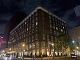 A large, multi-story hotel building dominates the scene, with illuminated windows and lights along the street. The structure has a rectangular, almost L-shape, with a dark brick facade. Several people can be seen walking on the sidewalks, and a few cars are visible on the road. The sky is dark with clouds, indicating it is nighttime. Streetlights and traffic signals add to the urban atmosphere.