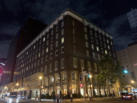 A large, multi-story hotel building dominates the scene, with illuminated windows and lights along the street. The structure has a rectangular, almost L-shape, with a dark brick facade. Several people can be seen walking on the sidewalks, and a few cars are visible on the road. The sky is dark with clouds, indicating it is nighttime. Streetlights and traffic signals add to the urban atmosphere.