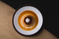 A round caramel custard dessert sits on a white plate, elegantly topped with a small swirl of whipped cream. The dessert appears to be a flan surrounded by a pool of golden-brown caramel sauce, contrasting with the dark background and coarse-textured, earth-toned tablecloth.