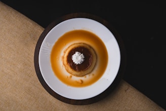 A round caramel custard dessert sits on a white plate, elegantly topped with a small swirl of whipped cream. The dessert appears to be a flan surrounded by a pool of golden-brown caramel sauce, contrasting with the dark background and coarse-textured, earth-toned tablecloth.