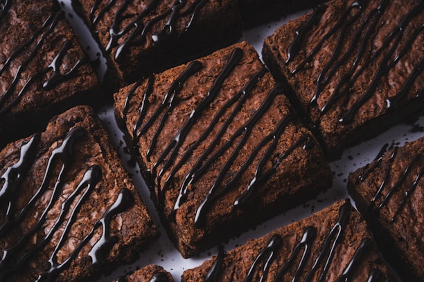 Several rich, fudgy brownies are arranged closely together. Each brownie is drizzled with a generous amount of glossy, dark chocolate syrup, creating an appetizing and indulgent appearance.