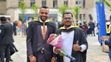 Two men wearing academic gowns and caps are standing outdoors during a graduation ceremony. One is holding a bouquet of pink flowers and a diploma holder. They are both smiling, and people in similar attire can be seen in the background, indicating a festive and celebratory atmosphere.