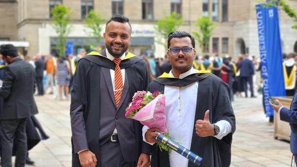 Two men wearing academic gowns and caps are standing outdoors during a graduation ceremony. One is holding a bouquet of pink flowers and a diploma holder. They are both smiling, and people in similar attire can be seen in the background, indicating a festive and celebratory atmosphere.