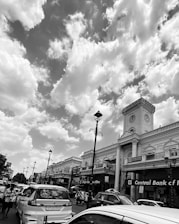 A busy street scene captured in black and white, featuring a historical building with clock towers and ornate architectural details. Cars are lined up along the road, indicating traffic or congestion. Street lamps line the sidewalk, and several commercial signs are visible, including one for the Central Bank of India. The sky is filled with large, fluffy clouds, adding a dramatic element to the scene.