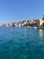 A picturesque coastal village with colorful buildings lining the waterfront. The vibrant blue sea occupies the foreground, while clear skies enhance the tranquil atmosphere. Small boats are docked along the pier, and the houses have red-tiled roofs and pastel walls.