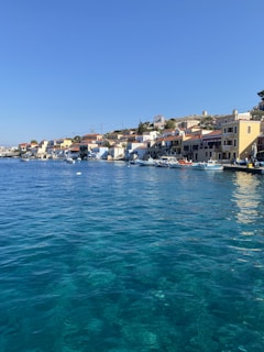 A picturesque coastal village with colorful buildings lining the waterfront. The vibrant blue sea occupies the foreground, while clear skies enhance the tranquil atmosphere. Small boats are docked along the pier, and the houses have red-tiled roofs and pastel walls.