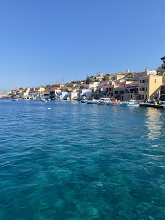 A picturesque coastal village with colorful buildings lining the waterfront. The vibrant blue sea occupies the foreground, while clear skies enhance the tranquil atmosphere. Small boats are docked along the pier, and the houses have red-tiled roofs and pastel walls.