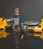 A perfume bottle labeled 'Black Afgano' stands upright surrounded by smooth black stones and two bright yellow flowers. The scene is reflected on a shiny black surface, and the bottle is decorated with water droplets.
