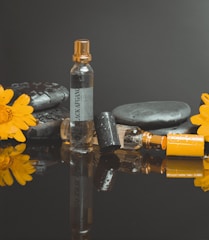 A perfume bottle labeled 'Black Afgano' stands upright surrounded by smooth black stones and two bright yellow flowers. The scene is reflected on a shiny black surface, and the bottle is decorated with water droplets.