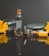 A perfume bottle labeled 'Black Afgano' stands upright surrounded by smooth black stones and two bright yellow flowers. The scene is reflected on a shiny black surface, and the bottle is decorated with water droplets.