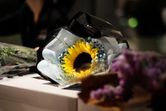 A beautifully wrapped bouquet featuring a prominent sunflower surrounded by small blue flowers and greenery. The bouquet is laid on a white surface and partially wrapped in black and white mesh. In the foreground, blurred purple flowers add a soft focus.