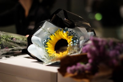 A beautifully wrapped bouquet featuring a prominent sunflower surrounded by small blue flowers and greenery. The bouquet is laid on a white surface and partially wrapped in black and white mesh. In the foreground, blurred purple flowers add a soft focus.