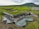 An architectural structure with a green roof seamlessly integrated into a grassy landscape. The building features organic-shaped pools and minimalistic design elements. Rolling hills and mountains are visible in the background.