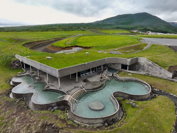 An architectural structure with a green roof seamlessly integrated into a grassy landscape. The building features organic-shaped pools and minimalistic design elements. Rolling hills and mountains are visible in the background.