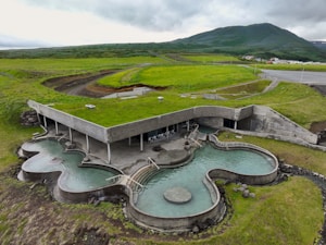An architectural structure with a green roof seamlessly integrated into a grassy landscape. The building features organic-shaped pools and minimalistic design elements. Rolling hills and mountains are visible in the background.