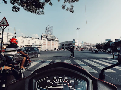 A busy urban intersection with vehicles and pedestrians. A person on a red scooter is wearing a red helmet and waiting at the crosswalk. The view from the handlebar reveals a speedometer. The background consists of a large, light-colored building with advertisements and clear blue sky.