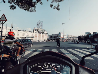 A busy urban intersection with vehicles and pedestrians. A person on a red scooter is wearing a red helmet and waiting at the crosswalk. The view from the handlebar reveals a speedometer. The background consists of a large, light-colored building with advertisements and clear blue sky.