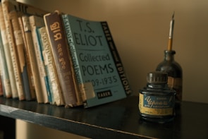 A shelf filled with a variety of books stands against a neutral wall. Prominently displayed is a blue book titled 'T.S. Eliot Collected Poems 1909-1935.' To the right of the books, there is an ink bottle with Russian text, accompanied by a quill pen resting inside a dark glass bottle.