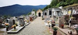 A serene cemetery with rows of headstones adorned with colorful flowers. The setting is surrounded by lush green mountains in the background, and there is a gravel pathway leading through the center. Tombs and mausoleums are visible along the sides, with some intricate stonework and statues.