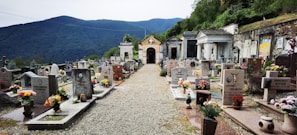 A serene cemetery with rows of headstones adorned with colorful flowers. The setting is surrounded by lush green mountains in the background, and there is a gravel pathway leading through the center. Tombs and mausoleums are visible along the sides, with some intricate stonework and statues.