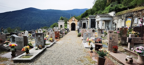 A serene cemetery with rows of headstones adorned with colorful flowers. The setting is surrounded by lush green mountains in the background, and there is a gravel pathway leading through the center. Tombs and mausoleums are visible along the sides, with some intricate stonework and statues.
